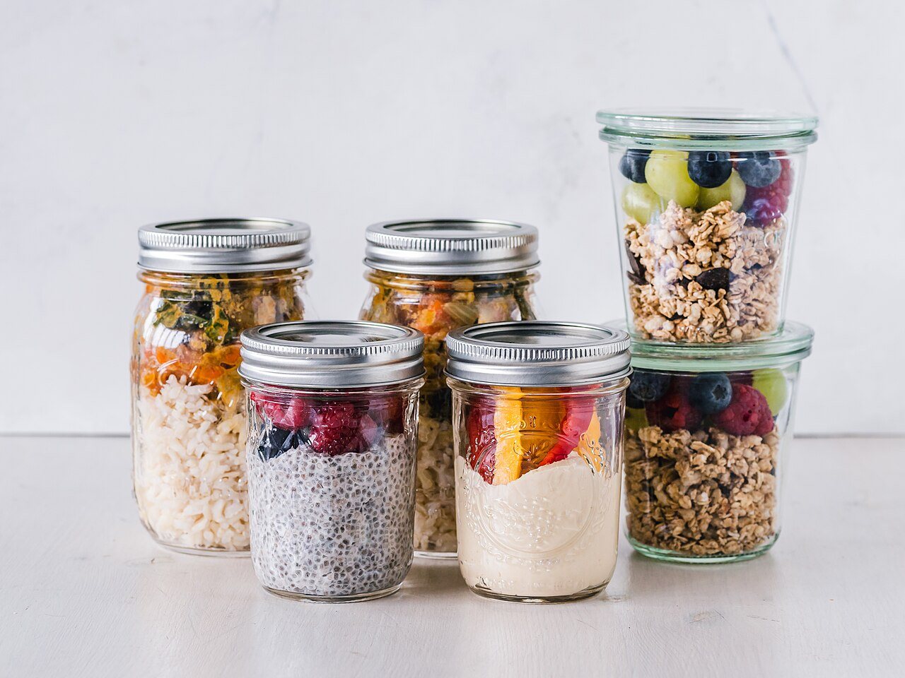 Glass jars of prepared meals on a white table: rice with vegetables, chia pudding with raspberries, yogurt with fruit and granola with berries